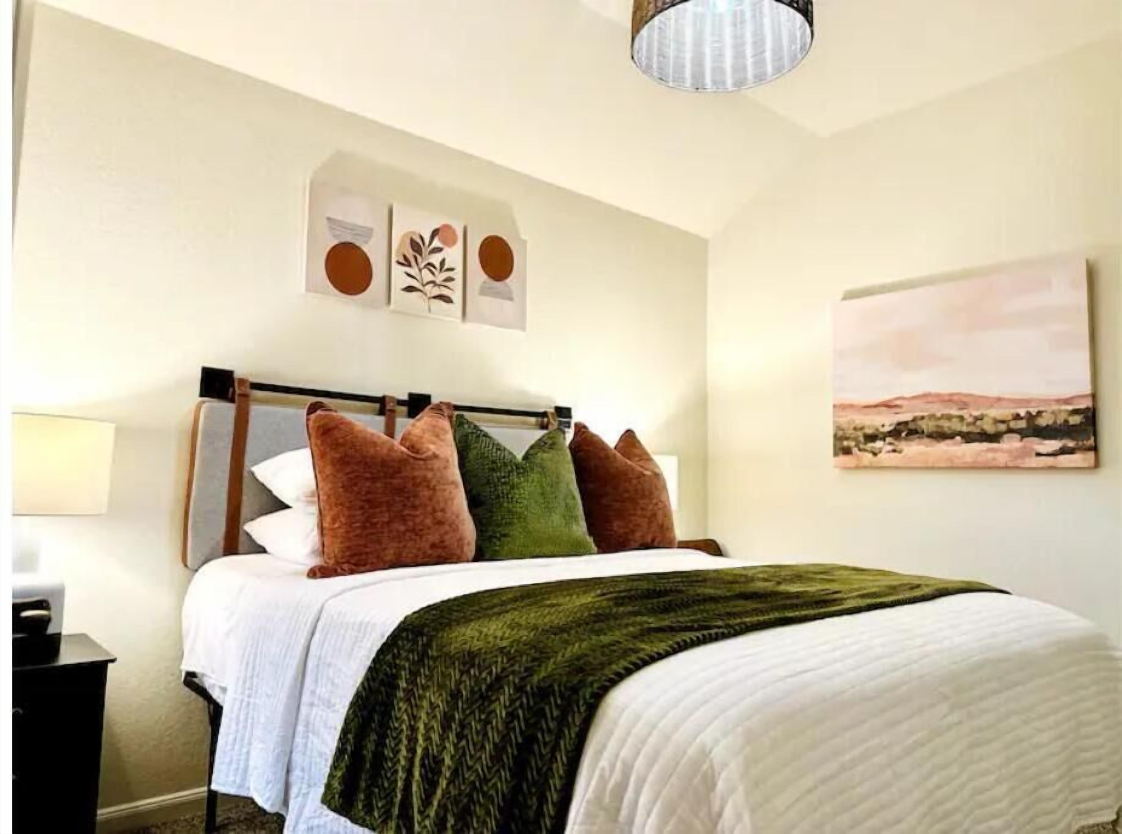 Cama bem arrumada com colcha branca e manta verde, cabeceira de madeira e abajur sobre a mesa de cabeceira.
