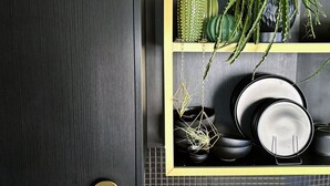A kitchen with a dark wood countertop, a yellow shelf with black and green plants, and black plates and bowls.