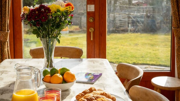 Une table de petit-déjeuner ornée d’un vase de fleurs, d’un bol d’oranges, d’une assiette de croissants, d’une carafe de jus d’orange et de verres à rayures.