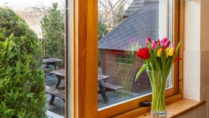Ein Fenster mit Holzrahmen und Scheibe, durch das ein Patio mit Holztisch und Stühlen sichtbar ist.