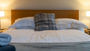 Una cama bien tendida con sábanas blancas, un cojín de cuadros y dos lámparas de noche.