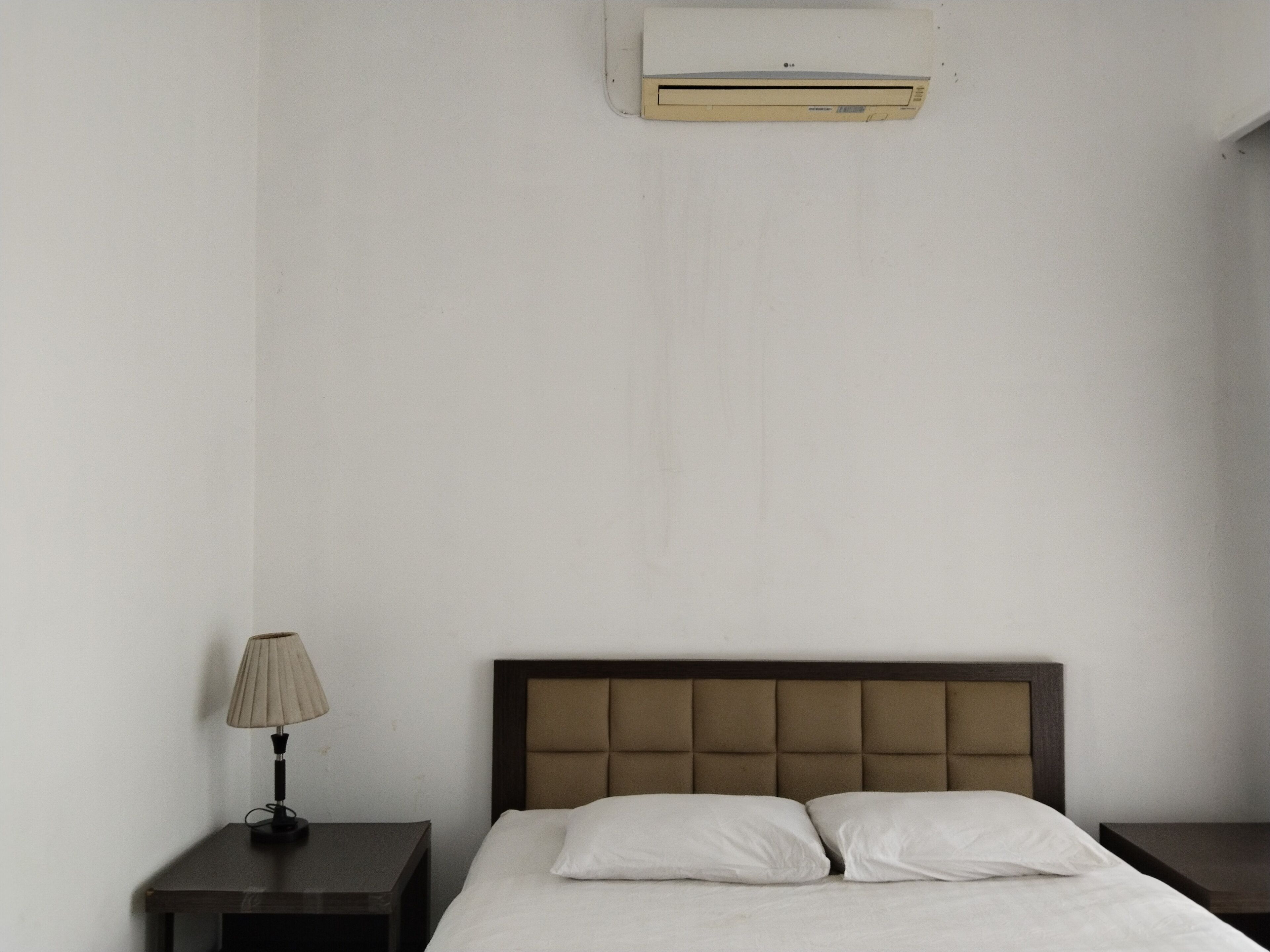 Ein schlichtes, sauberes Hotelzimmer mit einem Einzelbett, einem Nachttisch mit Lampe und einer Wand-Klimaanlage.