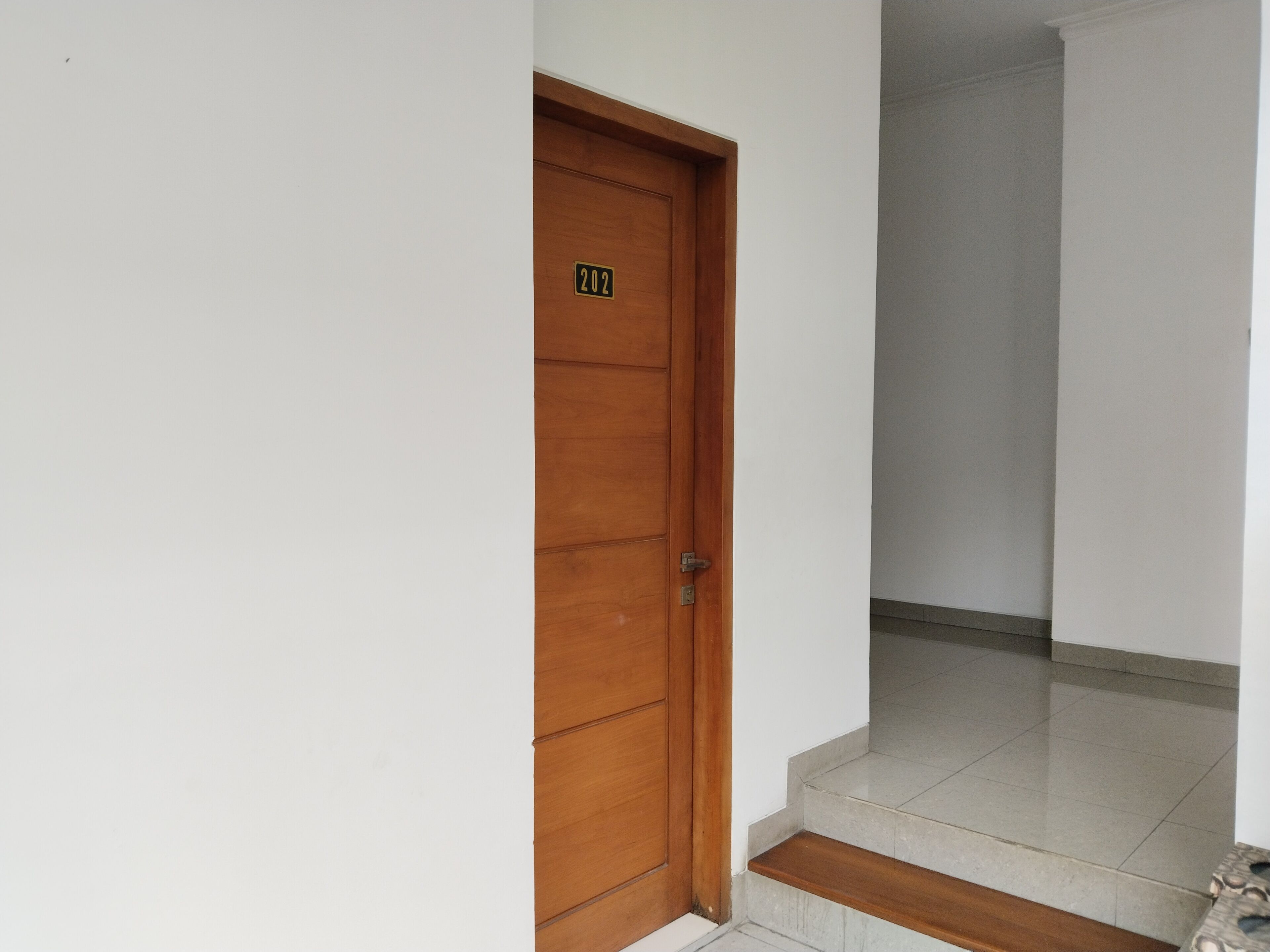 Puerta de madera con placa numérica, suelo de baldosas y una escalera que sube a un nivel superior.