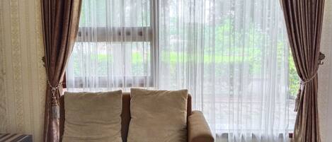 A room with a brown sofa, two beige cushions, and a large window with sheer white curtains.
