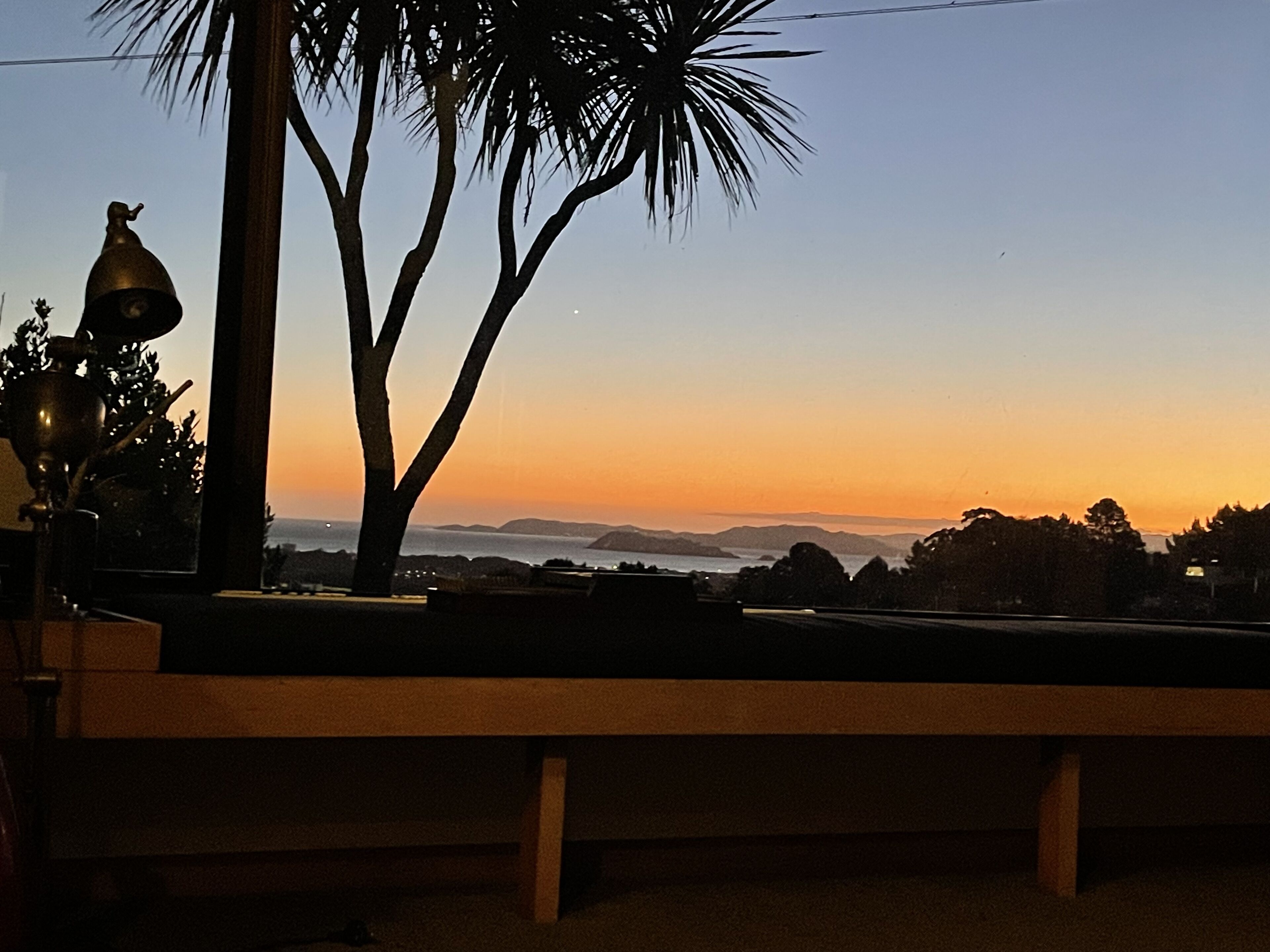 Zonsondergang vanaf een balkon met een bankje, een palmboom en een lamp.