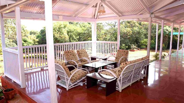 Un espace extérieur aménagé, abrité et doté de mobilier en osier blanc et d’une table en verre, le tout entouré d’une rambarde blanche, et se fondant dans un décor de verdure luxuriante.