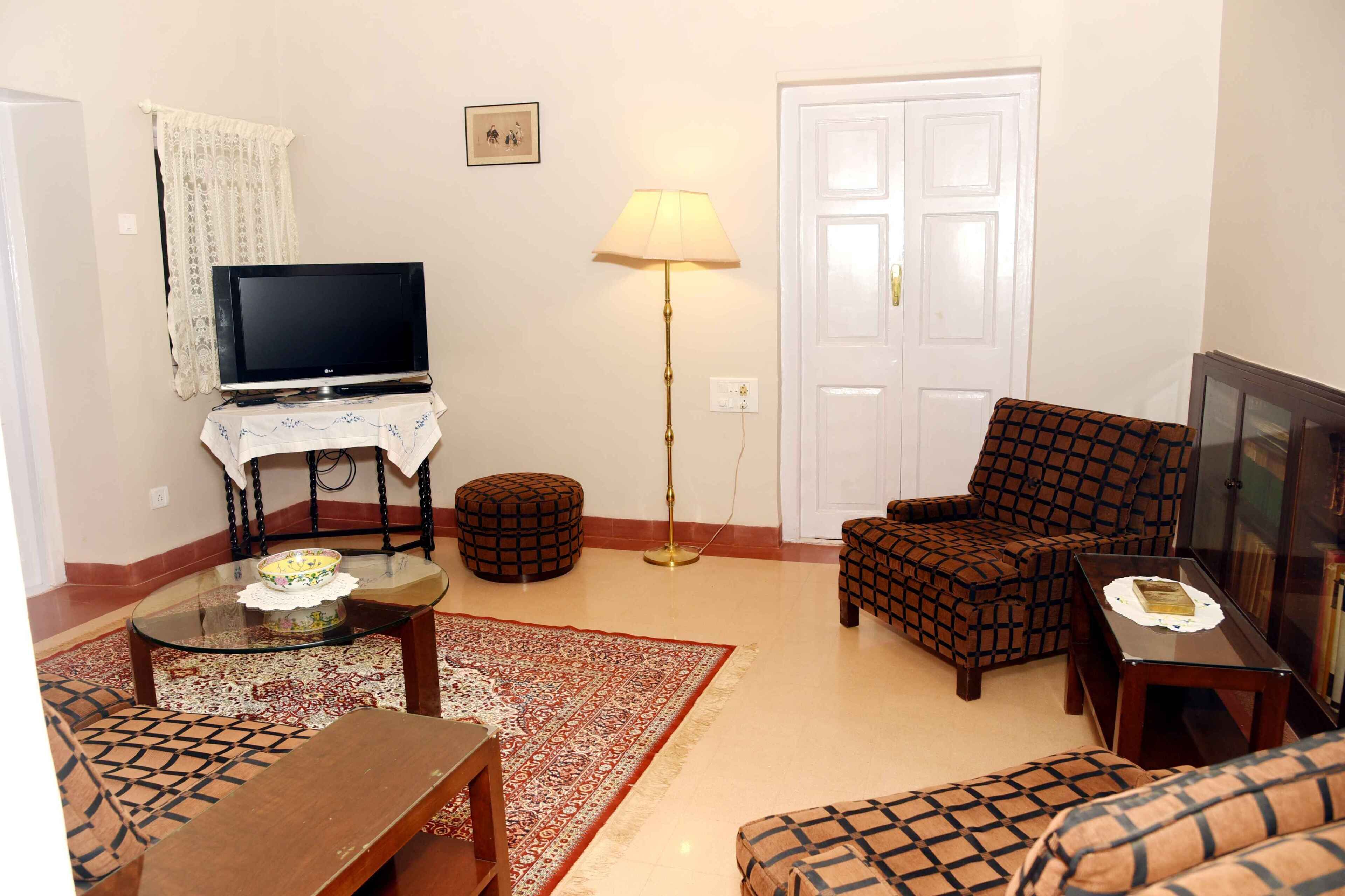 Una sala de estar con televisión, lámpara, sillón estampado, otomanes y una mesa de centro.