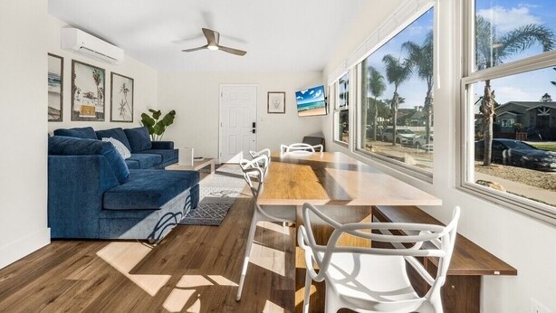 Un salon moderne avec un canapé bleu, des chaises blanches, une table à manger en bois et de grandes fenêtres donnant sur des palmiers et des maisons.