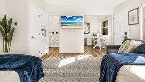 Une pièce moderne et bien éclairée, comprenant deux lits, un coin repas et une cuisine visible à travers une porte ouverte.