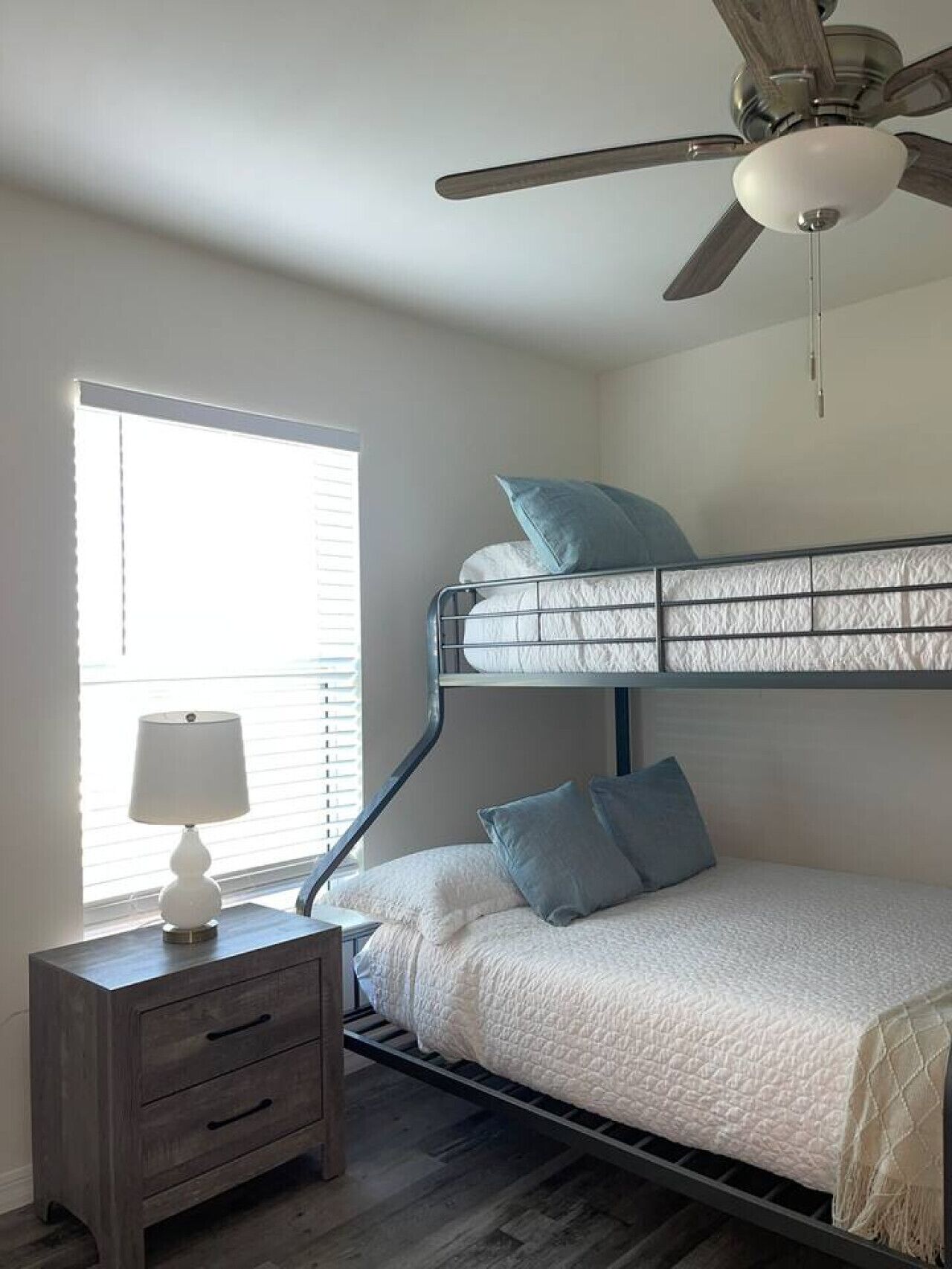A bunk bed with a ceiling fan, a bedside table with a lamp, and a window with blinds.
