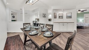 Un comedor con una mesa de madera dispuesta para cuatro personas, armarios blancos al fondo y una cocina visible a través de un umbral abierto.