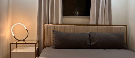 Quarto com cama, mesa de cabeceira com ring light, cortinas e janela.