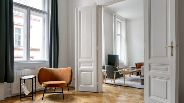 Ein geräumiges Zimmer mit Holzboden im Fischgrätmuster, einem braunen Sessel, einem kleinen Beistelltisch und Blick auf eine Stadtlandschaft durch große Fenster.