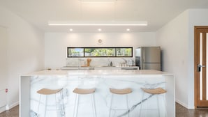 A modern kitchen with a marble island, bar stools, stainless steel appliances, and a view through the window. - Take it easy at this unique and tranquil getaway. (Tintenbar)