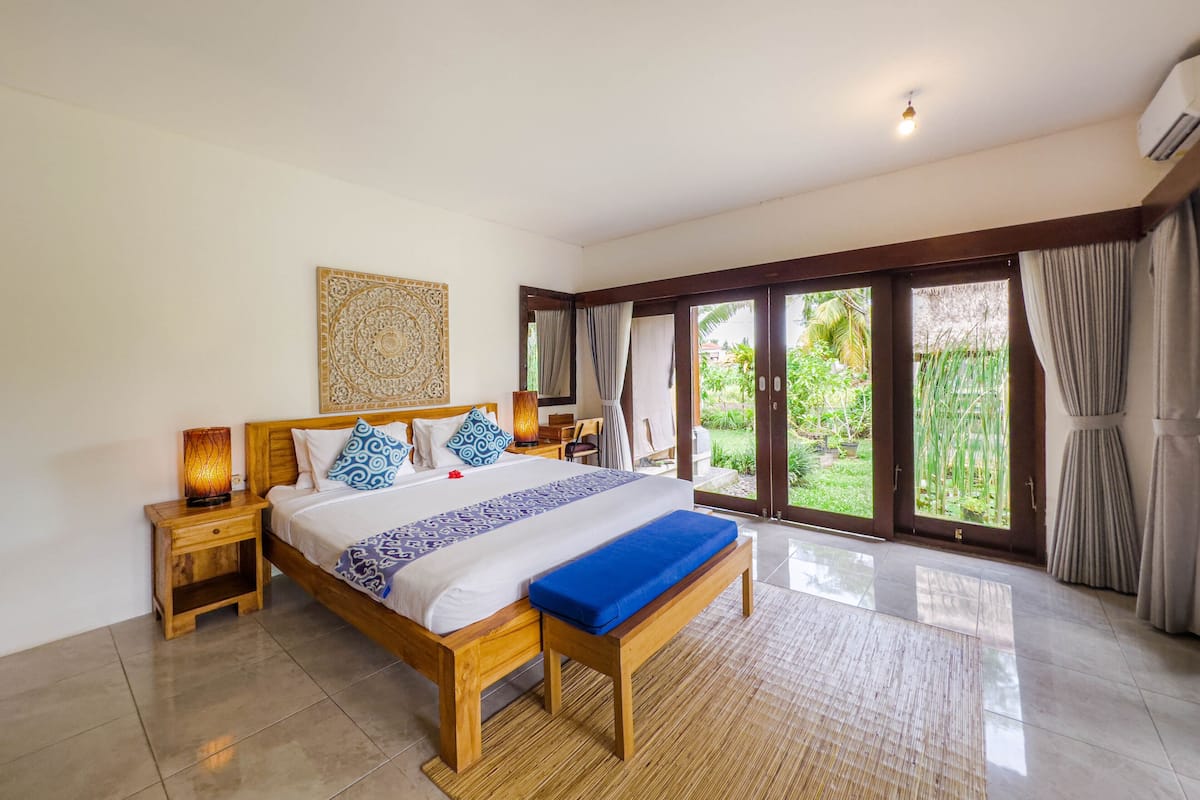 Une chambre spacieuse avec un grand lit, une tête de lit en bois et une vue sur un jardin, accessible par une porte-fenêtre coulissante.