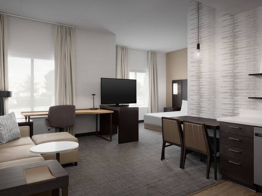 A modern hotel room with a sofa, a desk, a television, a bed, and a kitchen area.
