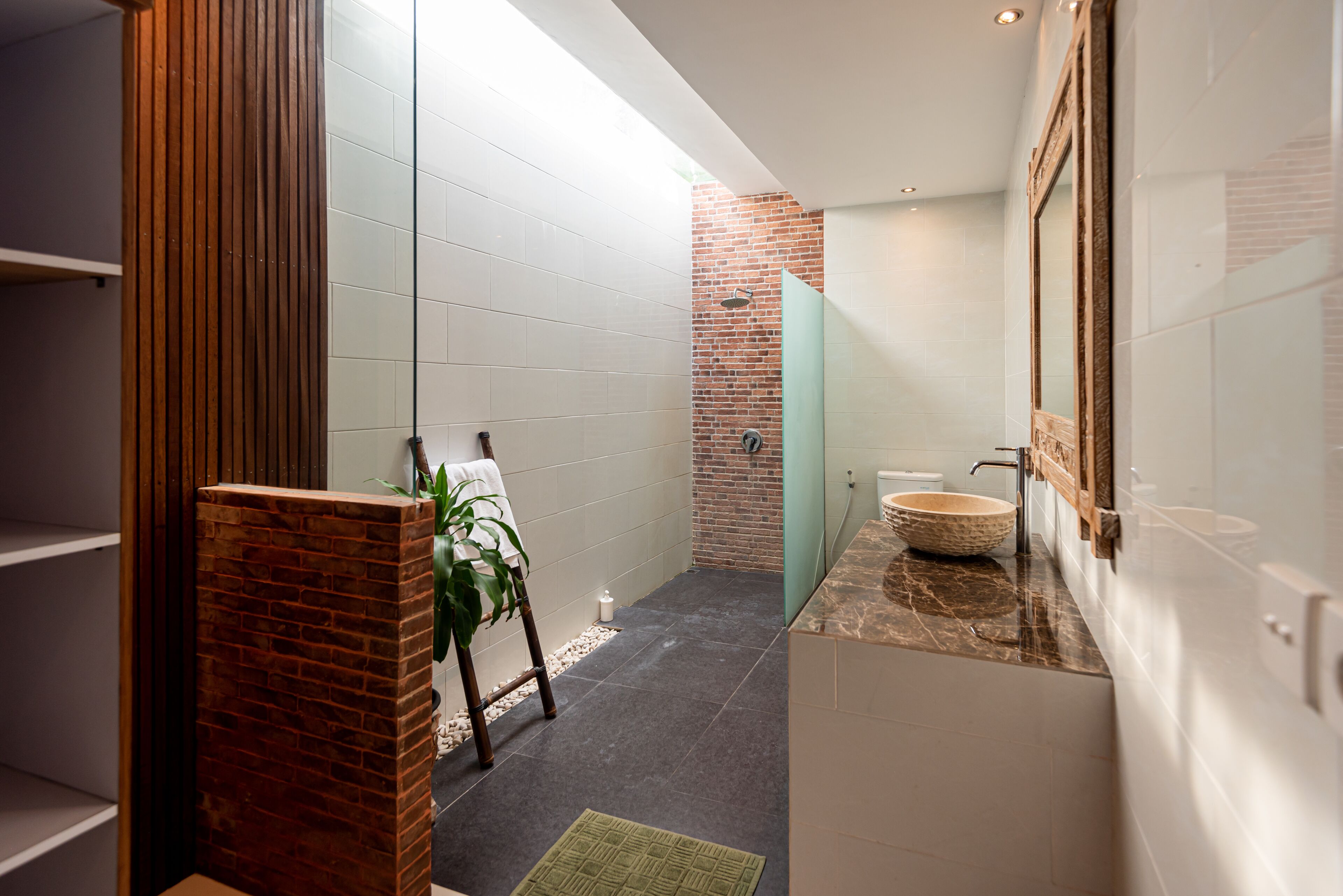 Une salle de bain moderne avec un mur d’accent en briques, une douche en verre, un lavabo en marbre et un support à plantes en bois.
