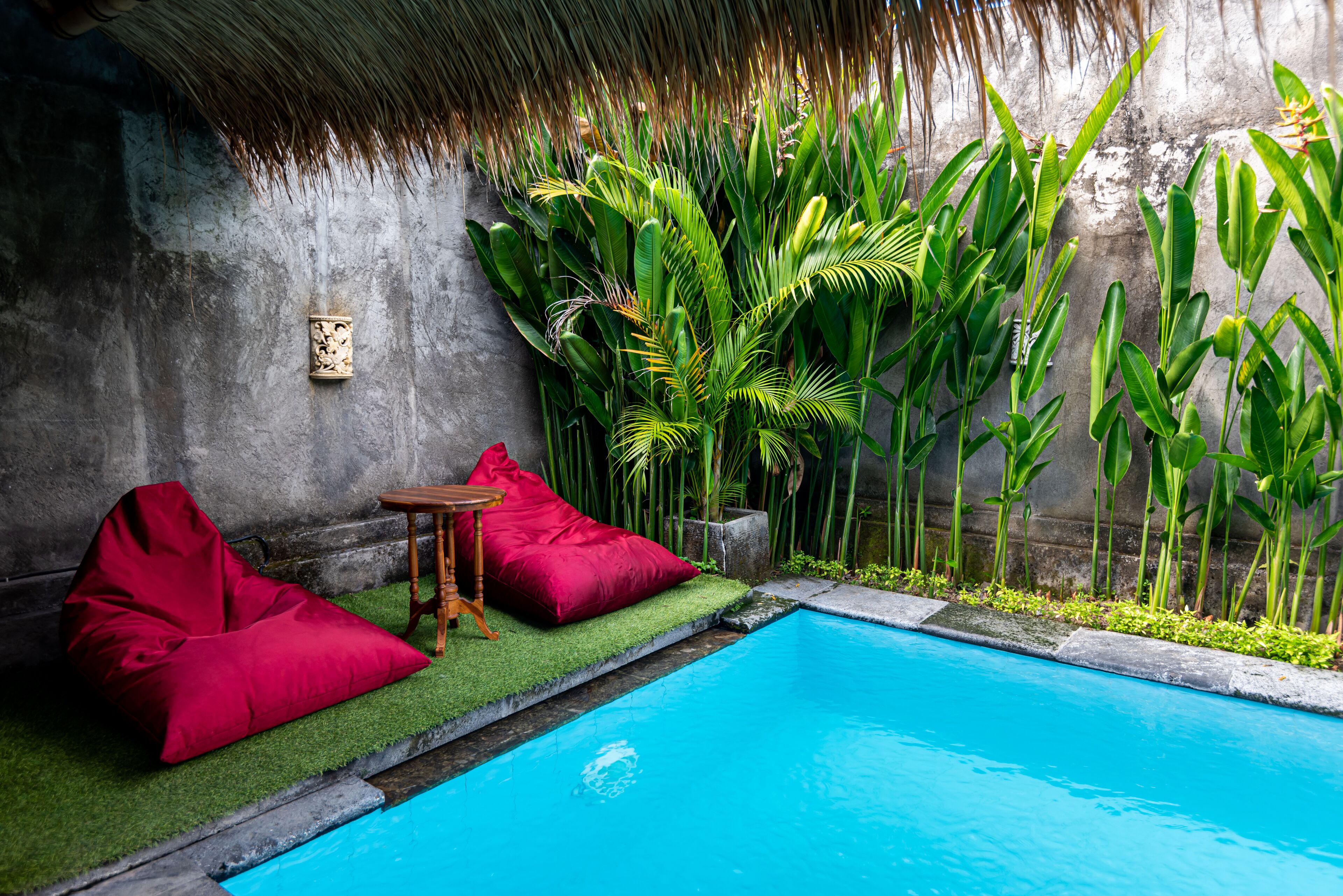 Un área de piscina con un sillón de mimbre rojo, un taburete de madera pequeño y abundante vegetación verde.
