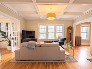Una sala de estar con un sofá color crema, un televisor, un reloj y un ventanal con vistas a edificios.