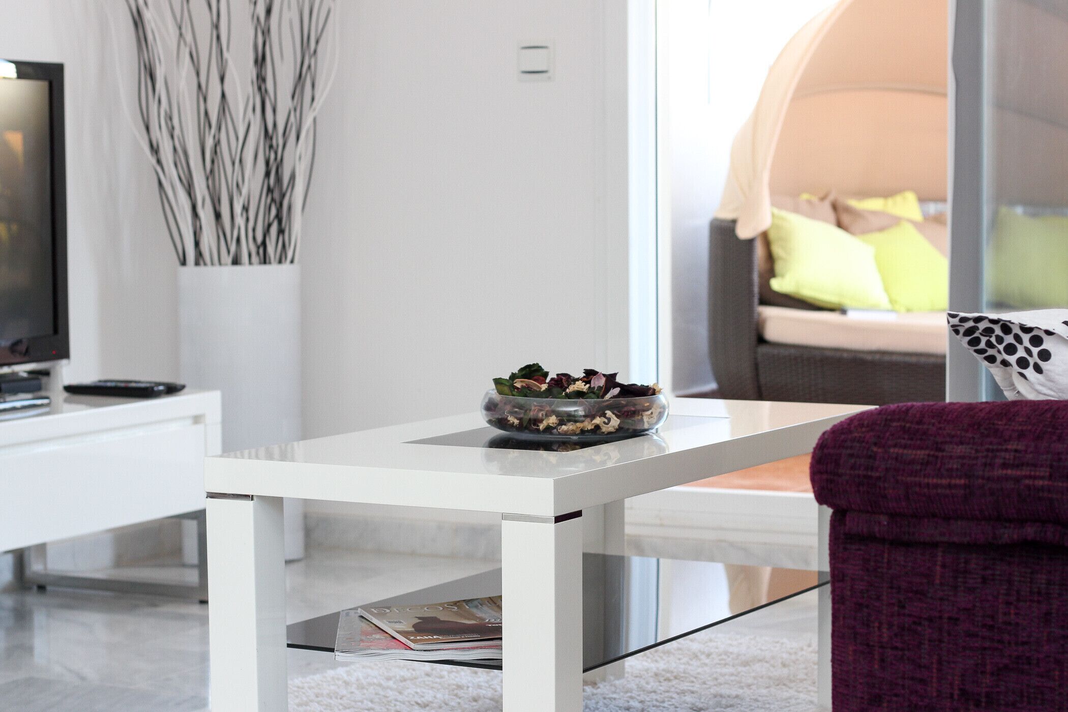 Una sala de estar moderna con una mesa blanca, una mesa de centro de vidrio, un sillón púrpura y un sofá con almohadas amarillas.