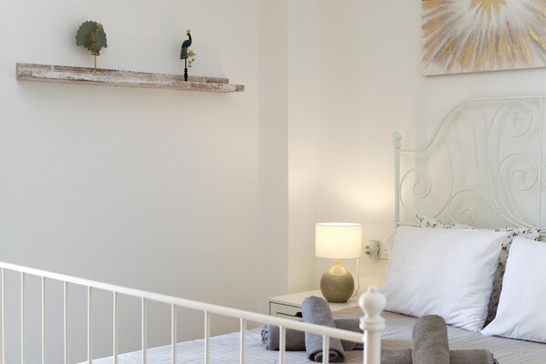 A bedroom with a white bed, a bedside table with a lamp, and decorative items on a shelf. - Cozy place in Fuengirola - 50m to the beach (Fuengirola)