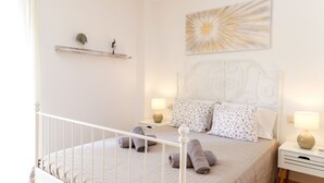 A neatly made bed with a headboard, two bedside tables with lamps, a wall-mounted shelf with decorative items, and a large abstract painting.