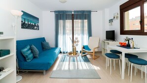 A modern living room with a blue sofa, a round coffee table, a white dining table with four chairs, a TV, and a painting on the wall.