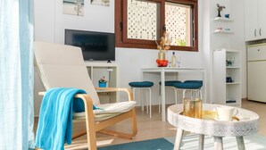 A living room with a TV, a blue-striped chair, a small table with a decorative vase, and a window with patterned curtains.