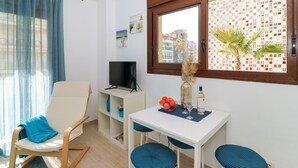 A modern living room with a white sofa, a small table with a vase and fruit, a TV, and a window with a view of buildings.