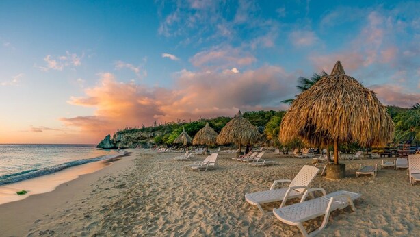 A beach with white lounge chairs and thatched umbrellas, a calm sea, and a sunset sky. - 08-Experience contemporary luxury at Prince APT, 20-minute stroll from the beach (Willemstad)