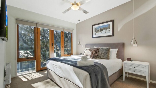 A bedroom with a large bed, a nightstand, a ceiling fan, a window with a view of trees, and a framed picture on the wall. - Apres - Dinner Plain (Dinner Plain)