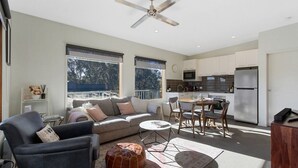 A modern living room with a sofa, armchair, and ottoman, a dining area with chairs, a kitchen with appliances, and large windows. - Apres - Dinner Plain (Dinner Plain)