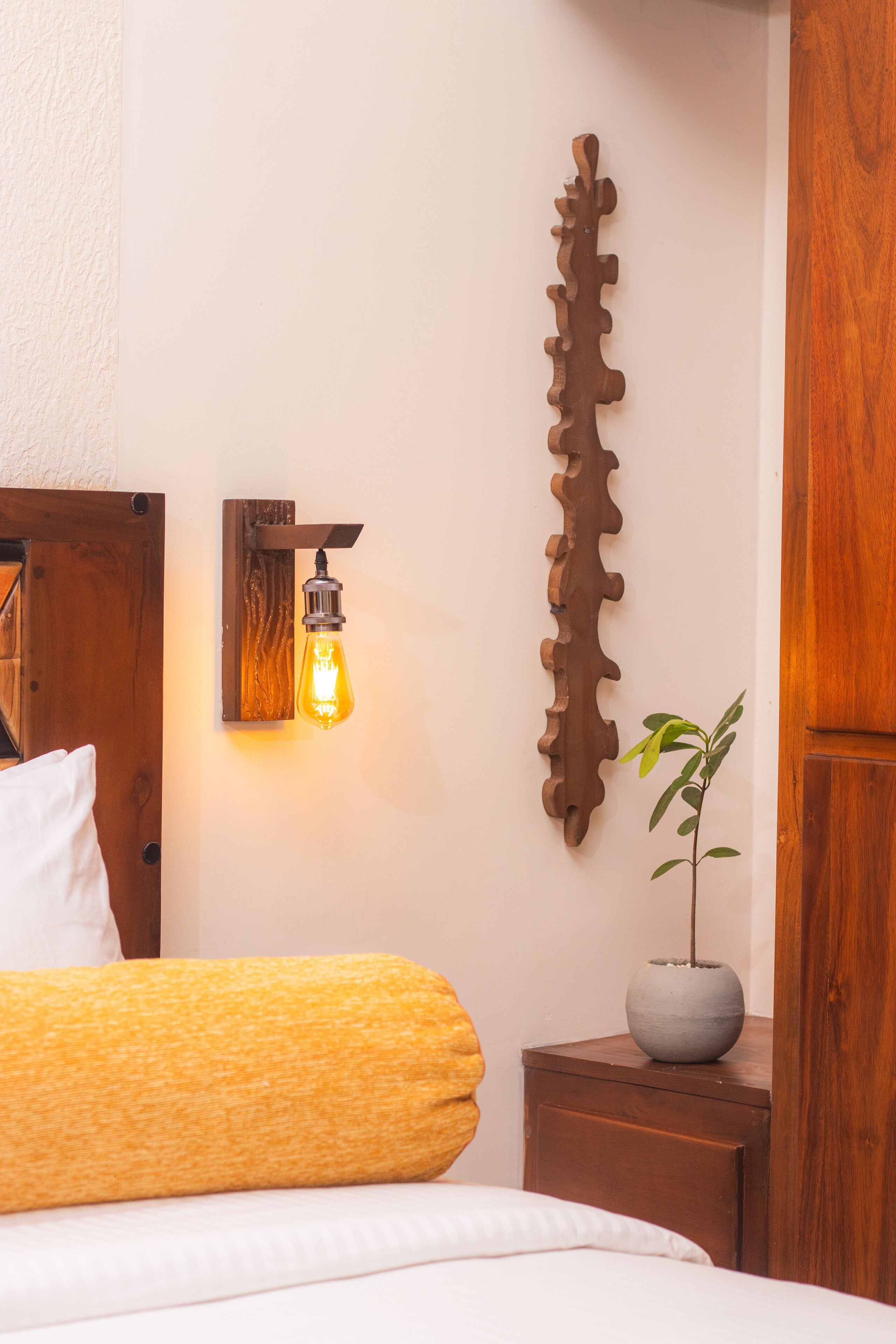 Ein ordentlich bezogenes Bett mit einem weißen Kissen, ein Nachttisch aus Holz mit einem Topf und Pflanze, eine Wandleuchte und ein dekoratives Holzschild an der Wand.
