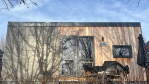 Een klein houten blokhutje met een metalen deur, een raam met uitzicht op bomen en een bordje waarop 