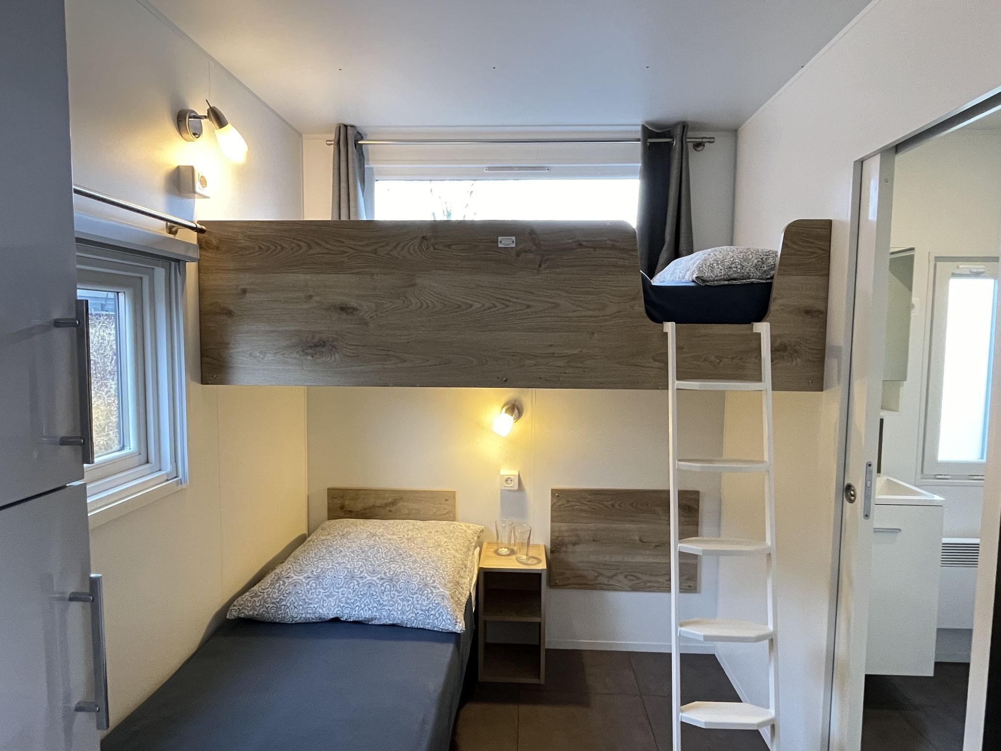 Une chambre compacte et moderne, équipée d’un lit superposé, d’un petit bureau avec une chaise, d’une fenêtre et d’une porte.