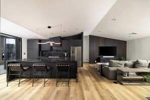 A modern kitchen with a central island, bar stools, a flat-screen TV, and a grey sofa in a living area.