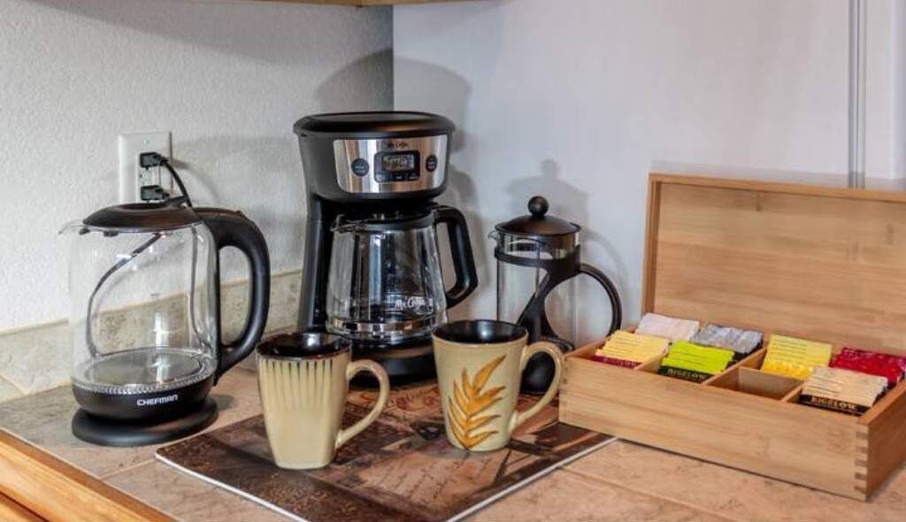 Una cocina con cafetera, jarra de vidrio, dos tazas y una caja de madera con bolsitas de té.