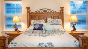 Un dormitorio con cabecera de madera, ropa de cama estampada, dos lámparas de noche y una ventana con persianas.