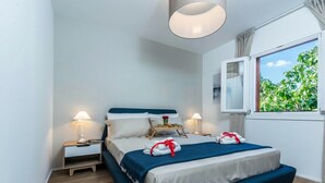 Ein modernes Schlafzimmer mit Bett, Nachttischlampen, einem Fenster mit Blick auf Grünflächen und einer Deckenleuchte.