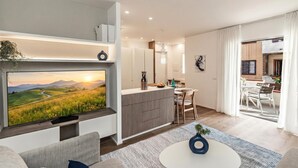 Ein modernes Wohnzimmer mit Flachbildfernseher, einer Couch, einem Couchtisch und einer Küchenzeile inklusive Esstisch und Stühlen.