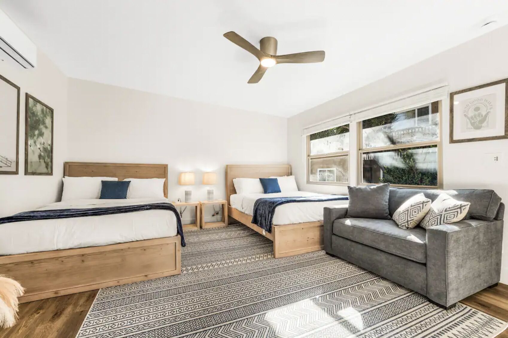 A hotel room with two beds, a grey sofa, a ceiling fan, and large windows.