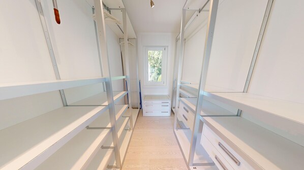 Un couloir étroit aux murs blancs, avec une fenêtre au fond. Des étagères bordent les deux côtés du couloir, et un radiateur est posé sur le sol.
