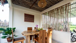A wooden dining table with chairs, a potted plant, and a decorative wall hanging in a bright room with a tiled floor and a woven ceiling. - Ulun Si Joglo Private Villa by Supala (Ubud)