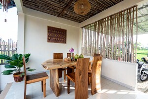 Una mesa de comedor de madera con sillas, una planta en maceta y un adorno en la pared en una habitación luminosa con suelo de baldosas y techo tejido.