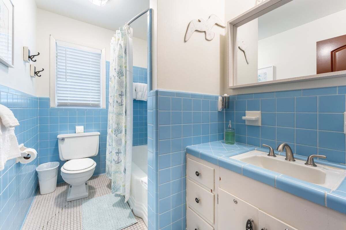 Un baño con azulejos azules, un inodoro blanco, un lavabo con dos grifos y un espejo sobre el lavabo.