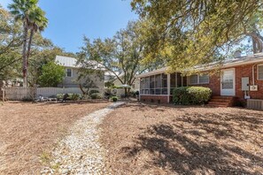 A residential property with a gravel driveway, a covered patio, and a brick building with large windows. - Family Tides | Pet-Friendly East Beach Cottage Near Gould's Inlet on St. Simons Island (Saint Simons Island)