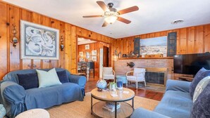 A living room with wooden paneling, a ceiling fan, a fireplace, and a television. - Family Tides | Pet-Friendly East Beach Cottage Near Gould's Inlet on St. Simons Island (Saint Simons Island)