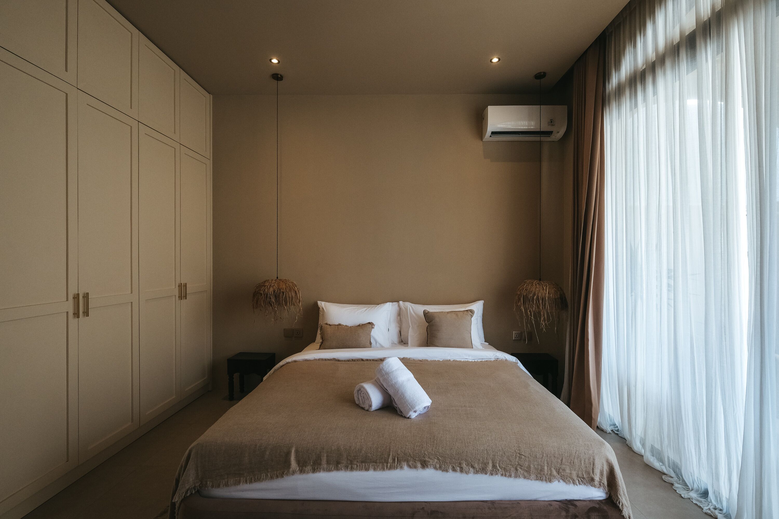 A bedroom with a large bed, a wardrobe, an air conditioning unit, and a window with sheer curtains.