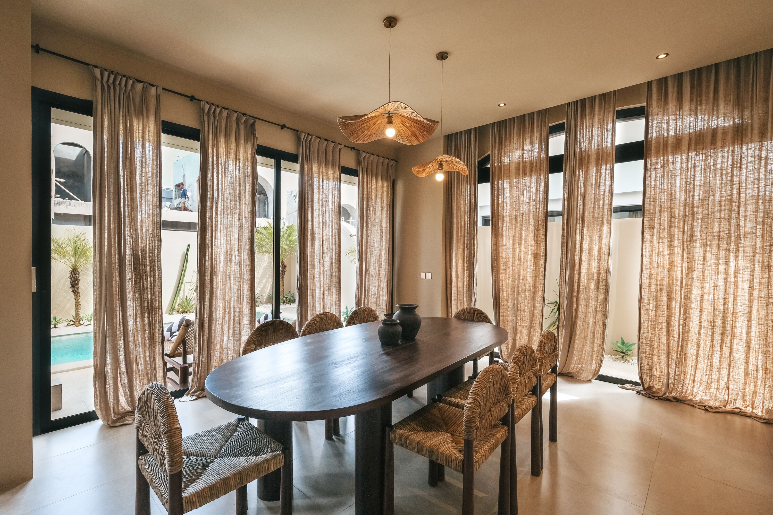 A dining area with a dark wooden table, woven chairs, and large windows with sheer curtains.