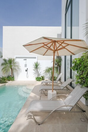 A modern outdoor pool area with lounge chairs and an umbrella, surrounded by greenery and a white building. - Limón & Cello Villa (Canggu)